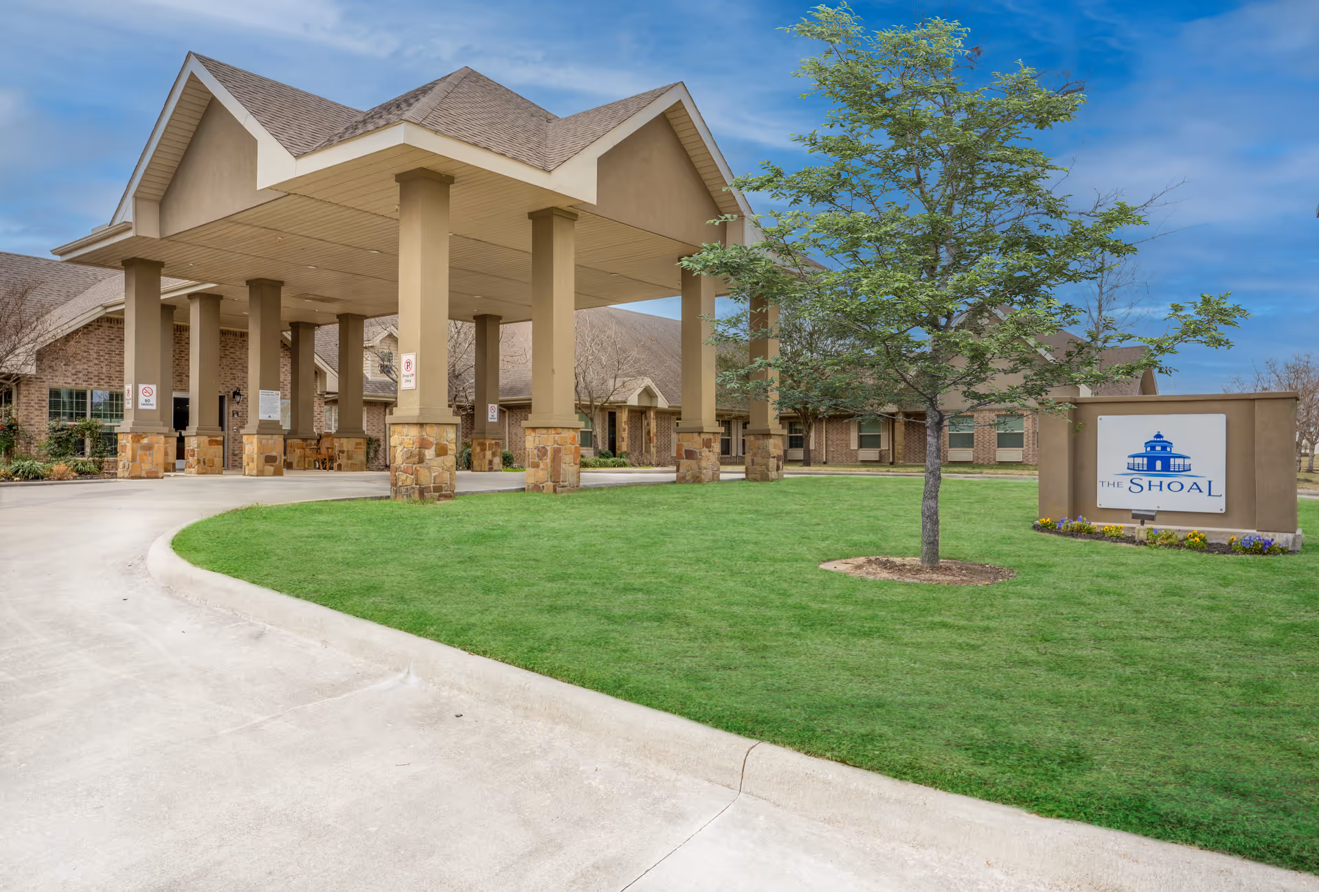 Exterior view of The Shoal senior living facility showing the entrance with a covered drop-off area supported by columns, a manicured lawn with a tree, and a sign displaying the facility's name and logo.