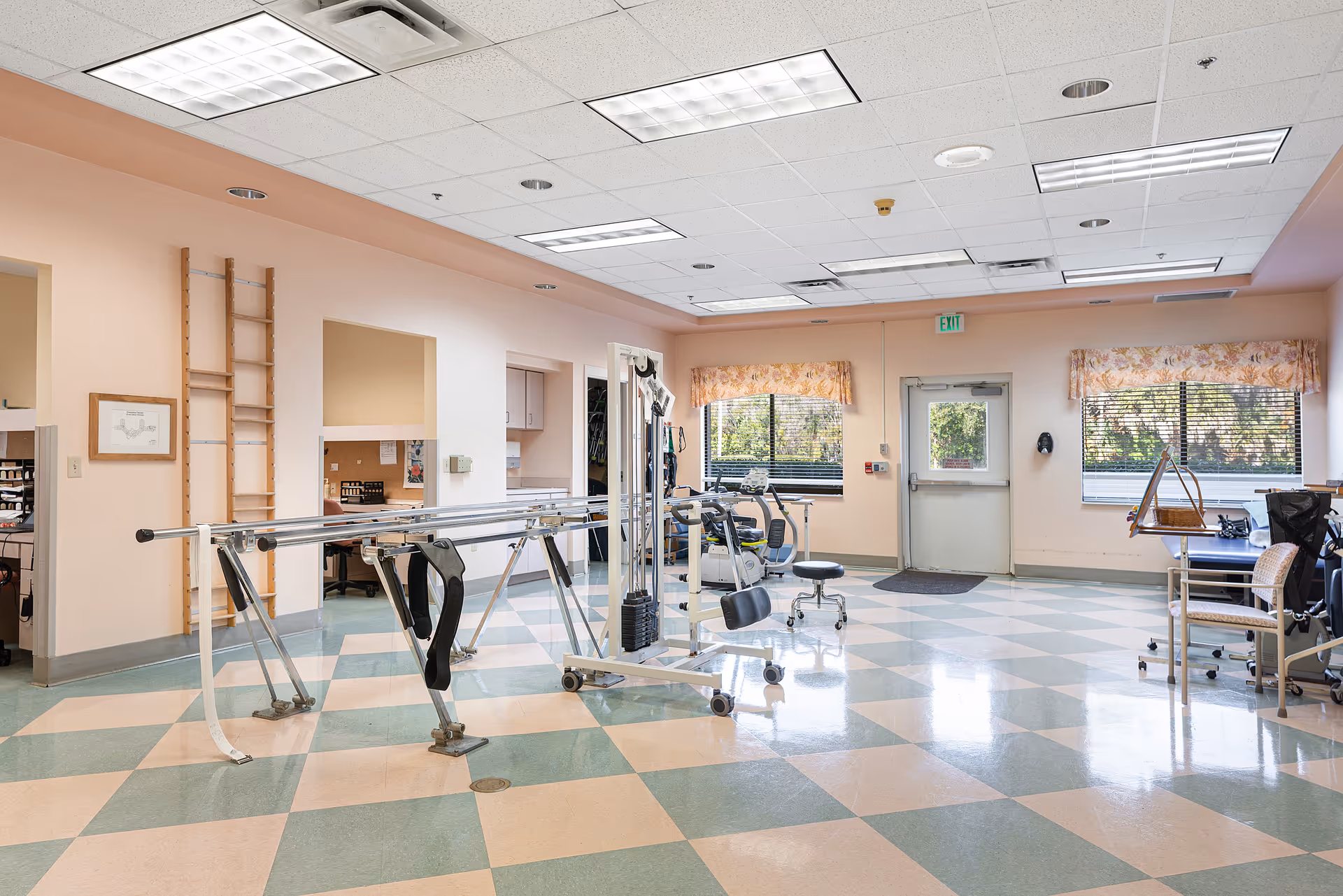 A spacious rehabilitation or physical therapy room with parallel bars for walking exercises, exercise equipment, chairs, and large windows with floral valances letting in natural light. The room has a checkered floor pattern in green and beige and peach-colored walls.