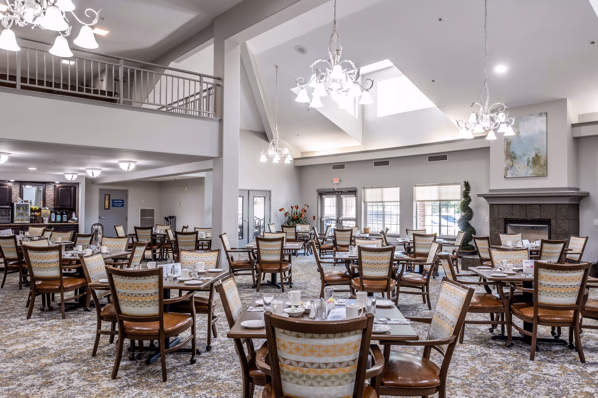 A spacious dining room in a senior living facility with multiple wooden tables and chairs arranged neatly. The room features high ceilings with hanging chandeliers, large windows letting in natural light, a fireplace with a painting above it, and a carpeted floor with a patterned design. The area appears clean and well-organized, ready for meal service.