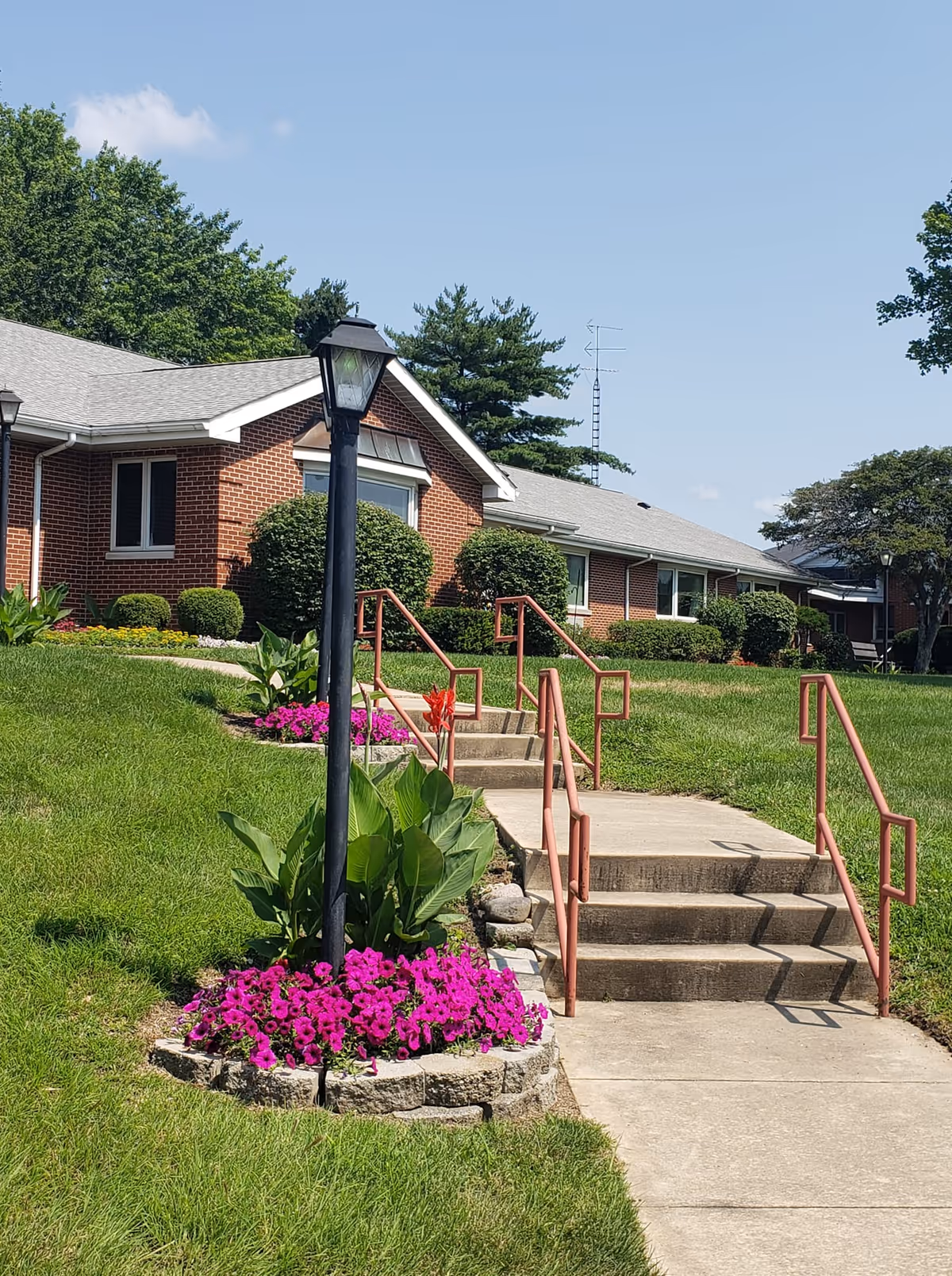 Outdoor view of a brick building with a gray roof, surrounded by green grass and landscaped flower beds with purple flowers. A concrete walkway with steps and red handrails leads up to the building. A black lamp post stands in the flower bed near the walkway. Trees and a clear blue sky are visible in the background.
