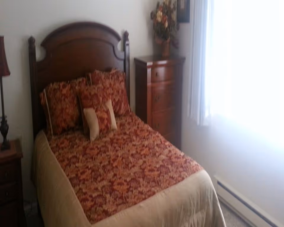 A small bedroom with a single bed featuring a dark wooden headboard and floral patterned bedding in shades of red and beige. There are matching pillows on the bed. To the right of the bed is a wooden chest of drawers with a flower arrangement on top. A window with white curtains allows natural light into the room.