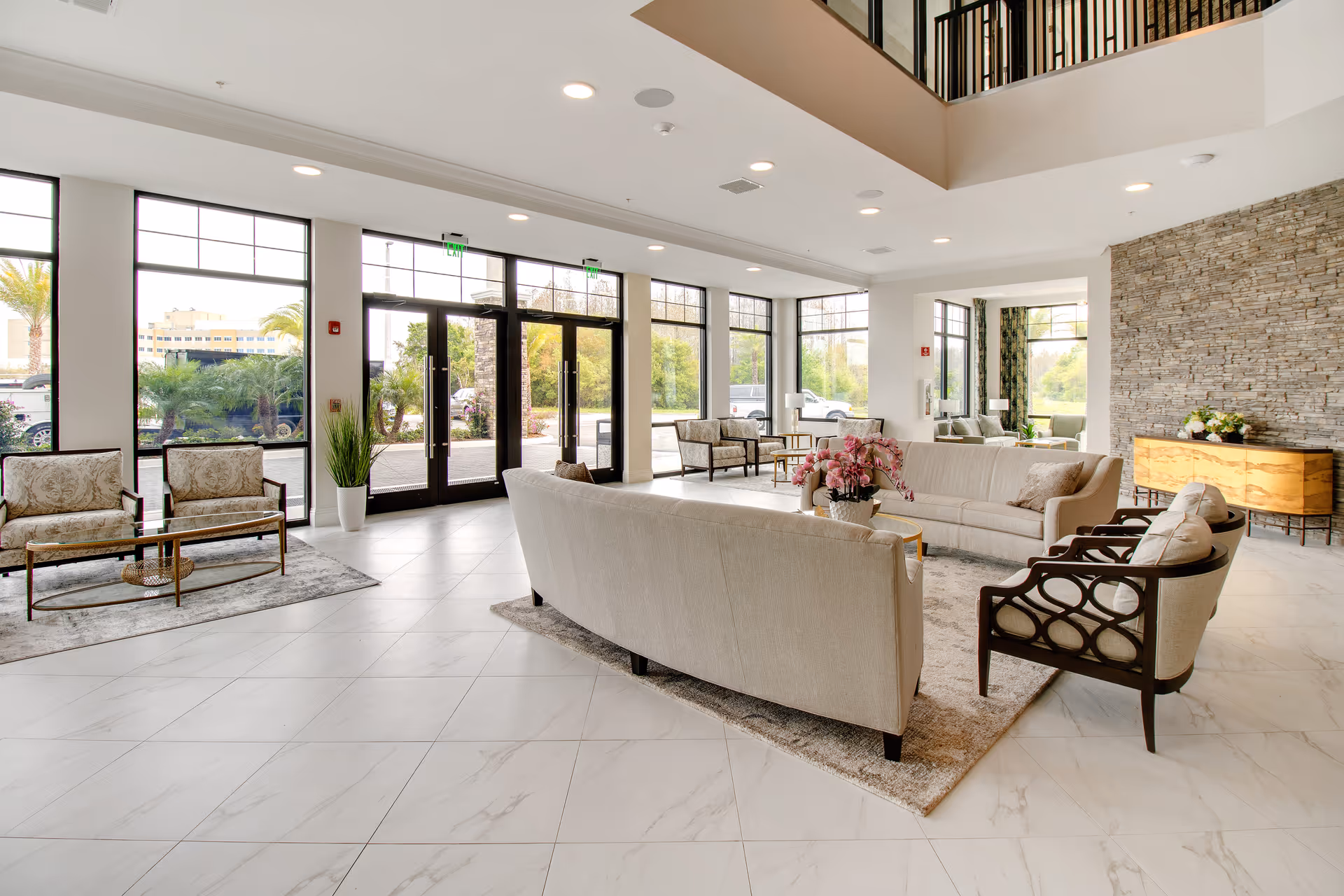 Bright and spacious assisted living facility lobby with large windows allowing natural light, beige sofas and armchairs arranged around a coffee table with a flower arrangement, a stone accent wall with a wooden console table, and tiled flooring.