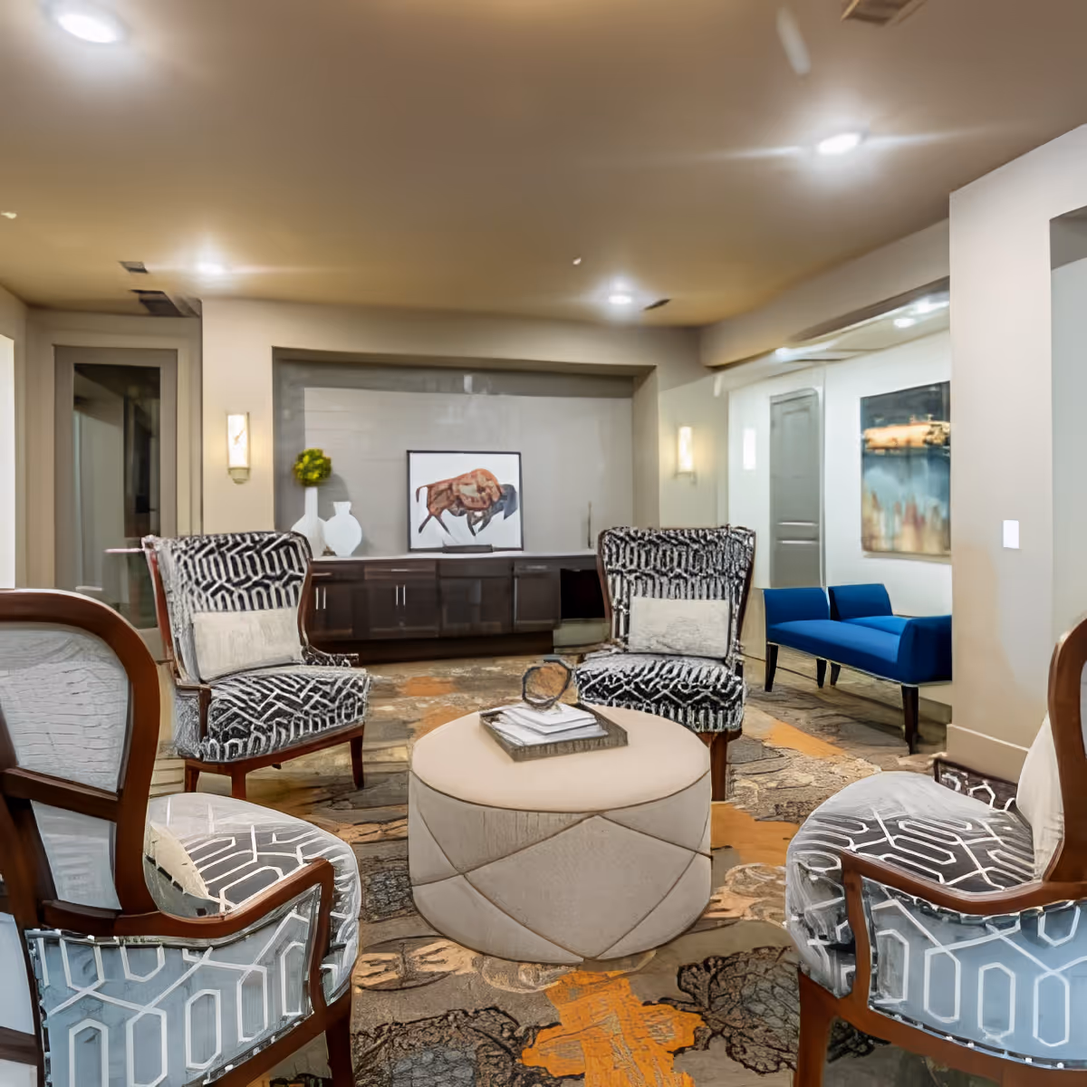 A cozy living room area with four patterned armchairs arranged around a round beige ottoman. The room features a decorative rug with orange and gray patterns, a dark wooden sideboard with vases and artwork above it, and a blue bench against the wall. The lighting is warm with ceiling lights and wall sconces.