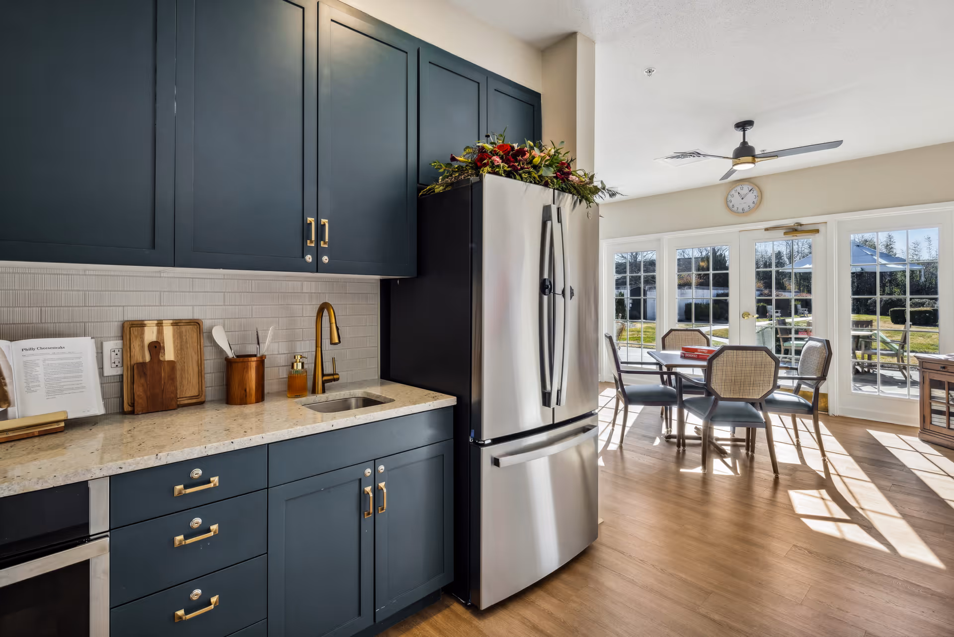 Bright modern kitchen with blue cabinets, a stainless-steel refrigerator and an adjoining sunlit dining area with a table and chairs.