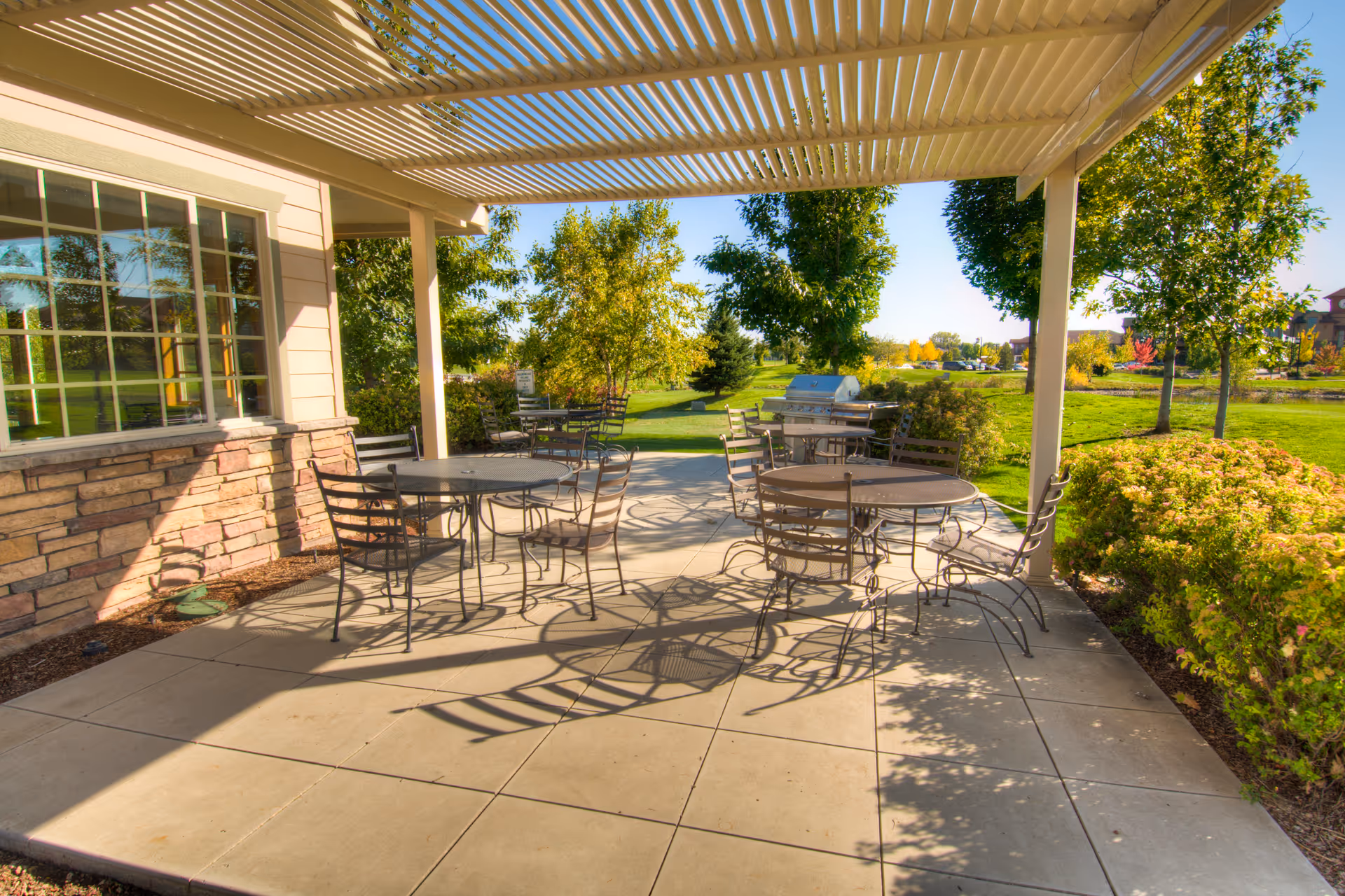 Outdoor patio area with metal tables and chairs under a pergola, surrounded by green trees and bushes with a grill in the background, adjacent to a building with stone and siding exterior.