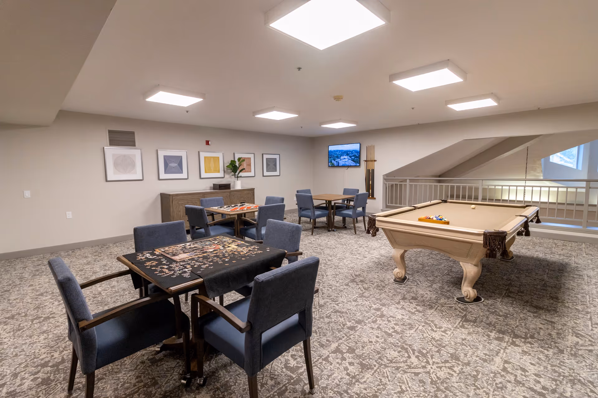A spacious recreational room with a pool table and several tables with chairs. One table has a partially completed jigsaw puzzle. The room has modern lighting, framed artwork on the walls, a TV mounted in the corner, and a rack holding pool cues. The carpet has a patterned design, and there is a railing overlooking a lower level.