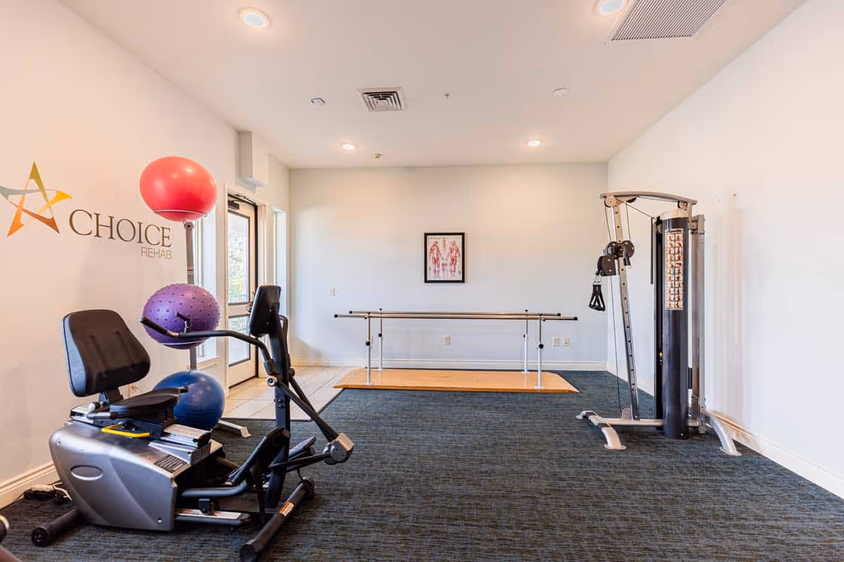 A rehabilitation exercise room with a recumbent exercise bike holding three colorful exercise balls, a parallel bar for walking practice, and a cable weight machine. The room has white walls, a dark carpeted floor, and a framed anatomical poster on the back wall. The left wall features the Choice Rehab logo.