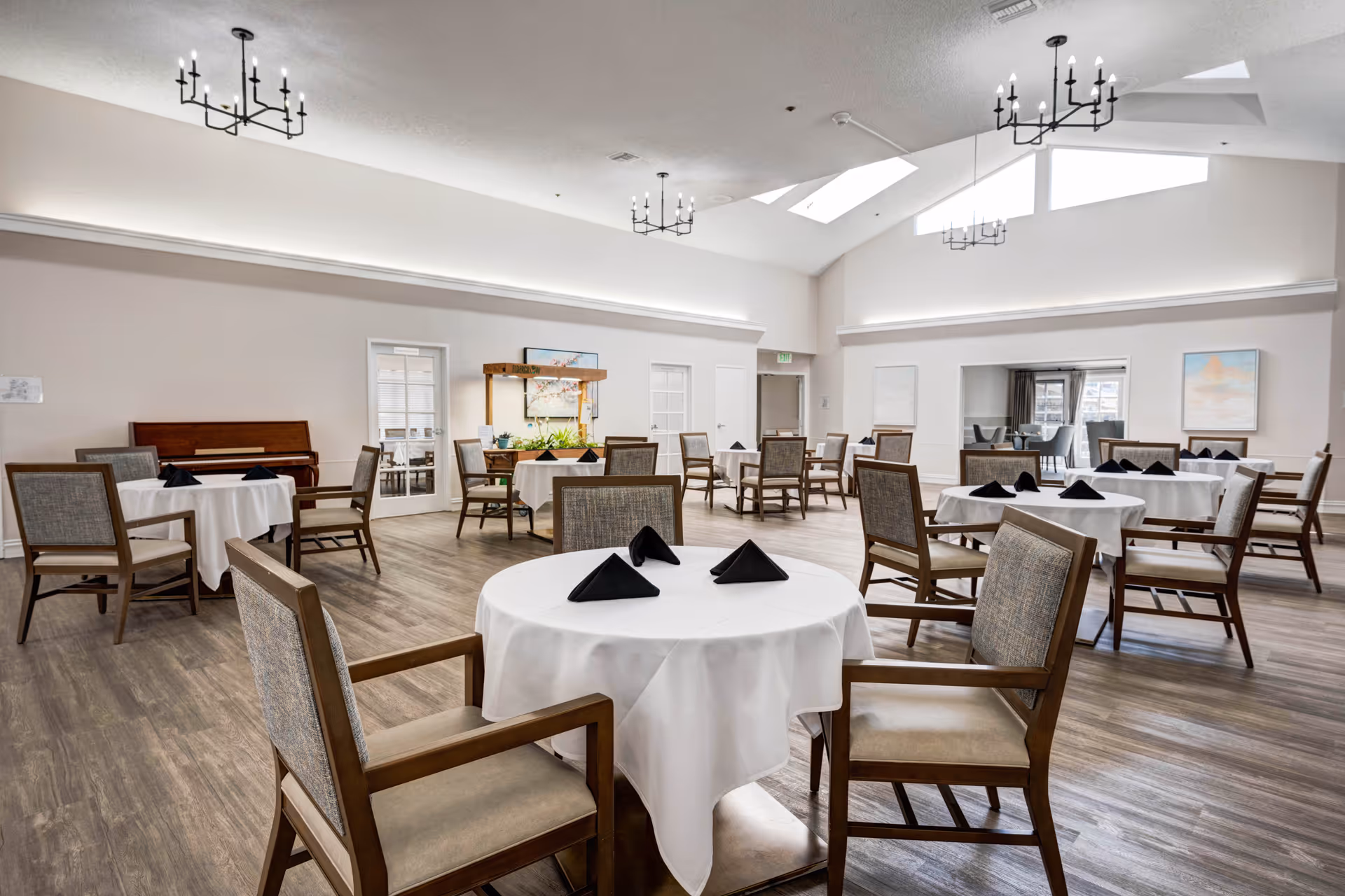 A spacious dining room with round tables covered in white tablecloths and set with black folded napkins. The room features wooden chairs with cushioned seats, wood flooring, and multiple modern chandeliers hanging from a high ceiling with skylights. There is a piano against one wall and a serving station with plants and artwork in the background.