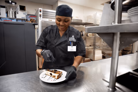 A woman wearing a black chef uniform and gloves is smiling while placing a fork on a slice of cake on a white plate in a commercial kitchen setting.
