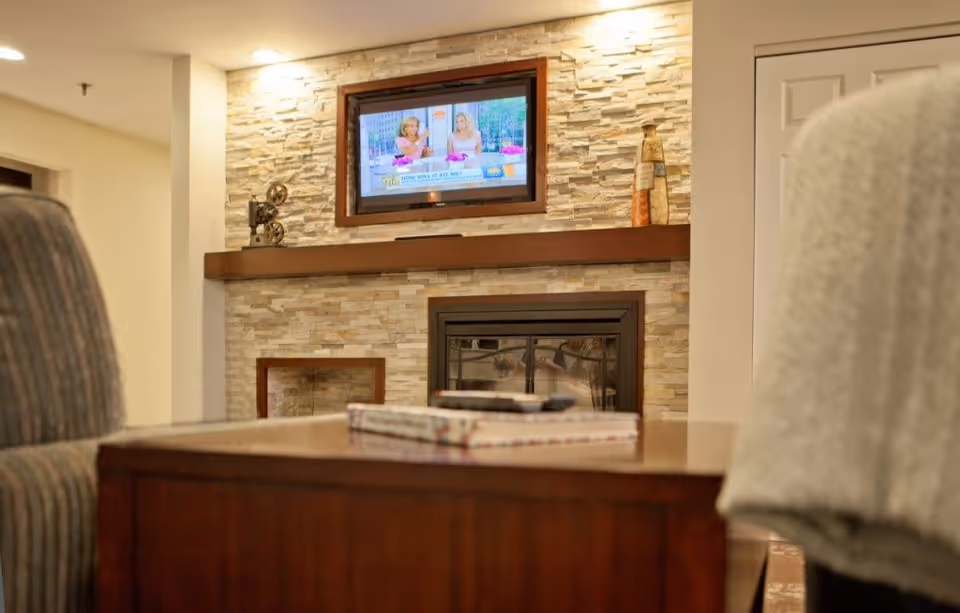 Cozy living room area with a stone fireplace, a mounted flat-screen TV above the mantel, and a wooden coffee table in the foreground. There are two chairs partially visible on either side of the table, one with a light-colored blanket draped over it.