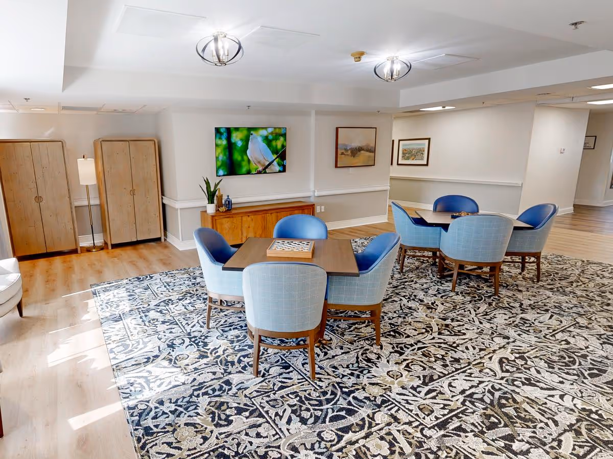 A bright and spacious common area with two square tables, each surrounded by four blue upholstered chairs. The floor features a large patterned rug, and the walls are decorated with framed artwork and a flat-screen TV displaying a bird. There are two wooden cabinets and a floor lamp in the corner, with light wood flooring and white walls.