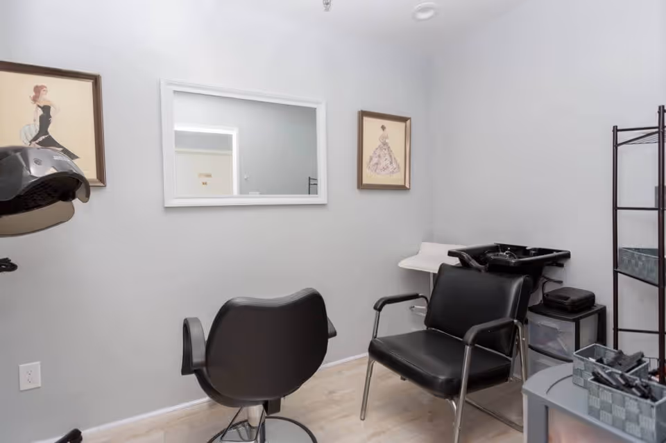 A small hair salon room with two black salon chairs, a hair washing station, a large white-framed mirror on the wall, two framed fashion illustrations, and a shelving unit with hair styling tools and supplies.