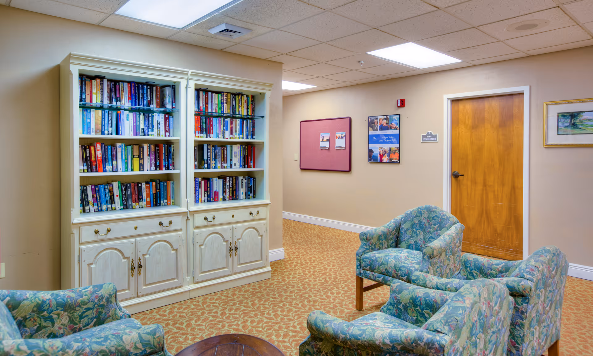 A cozy reading or sitting area in a senior living facility featuring a white bookshelf filled with books, four floral-patterned armchairs arranged around a small wooden table, beige walls with a bulletin board and framed pictures, and a wooden door labeled 'Live Enrichment'.