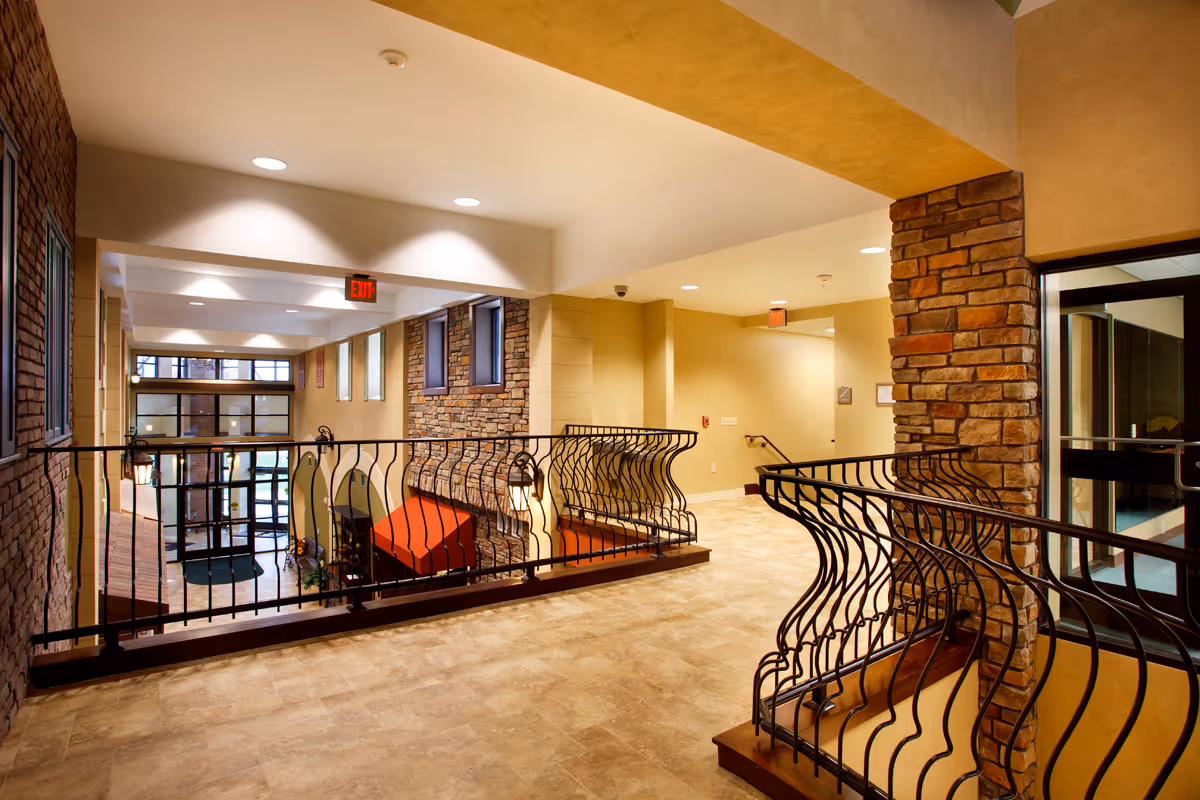 Second-floor interior corridor with decorative wrought-iron railings, stone columns, and a view down to a lobby area with seating and large windows.