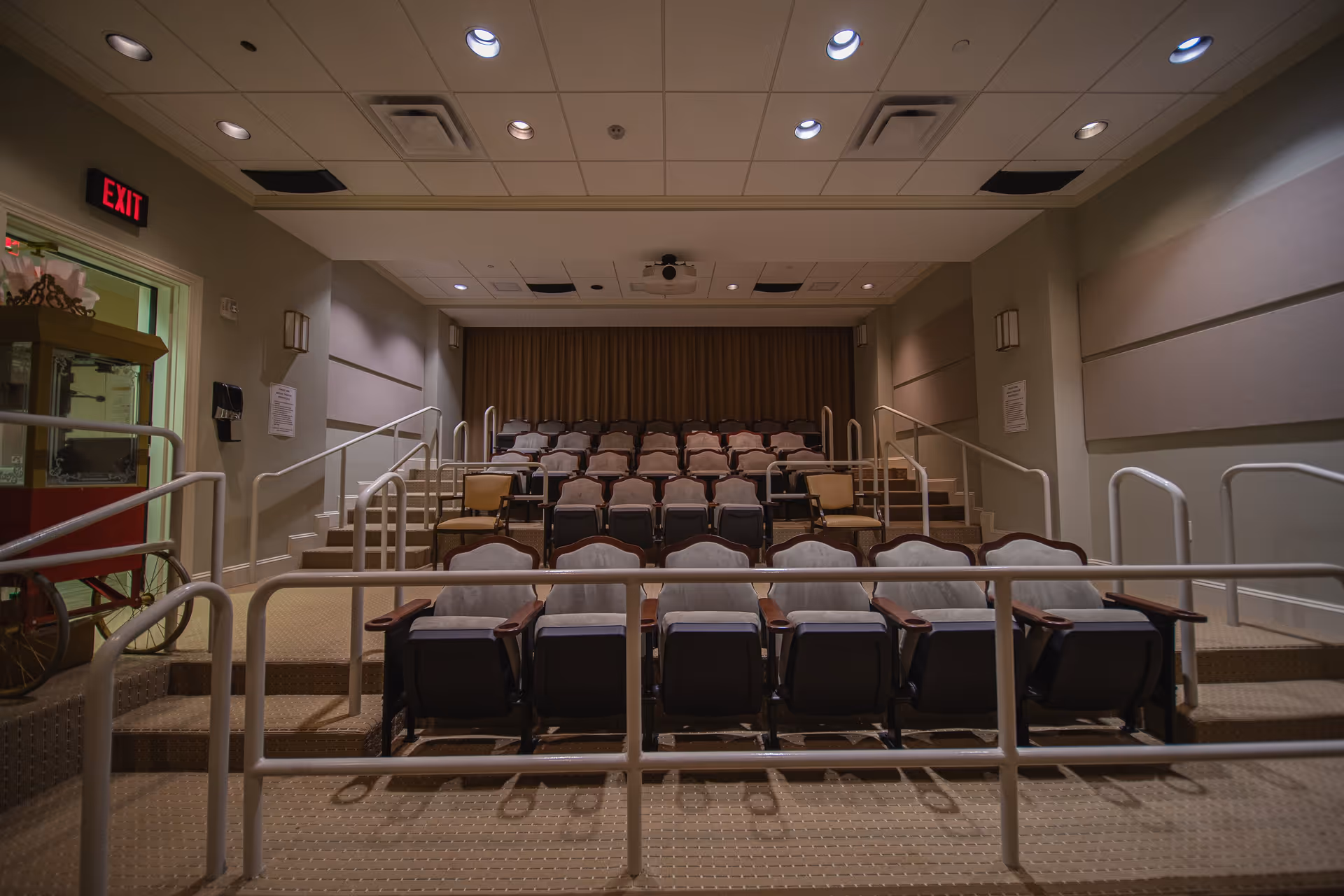 A small auditorium or theater room with tiered seating, including cushioned chairs and a few armchairs. The room has beige walls, a carpeted floor, handrails along the steps, and a projector mounted on the ceiling. There is an exit sign illuminated in red on the left side near a popcorn machine.