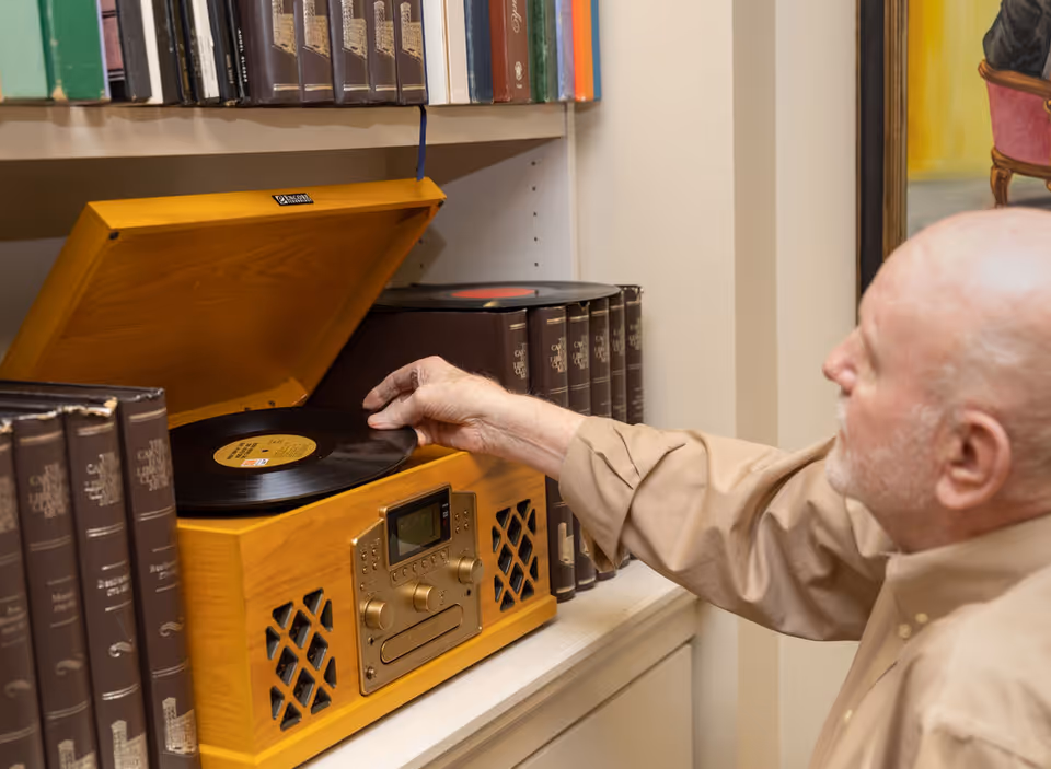 An elderly man placing a vinyl record on a wooden record player in a room with shelves filled with books.