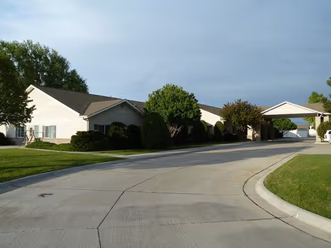 Exterior view of a single-story assisted living facility building with beige siding, surrounded by green grass, bushes, and trees under a cloudy sky. A paved driveway curves in front of the building leading to a covered entrance.