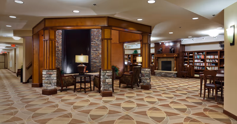 A spacious and well-lit common area in a senior living facility featuring a patterned carpet, wooden pillars with stone bases, comfortable armchairs around a small table with a lamp, a fireplace with a stone hearth, and bookshelves filled with books. There are additional seating areas with chairs and tables, and the ceiling has recessed lighting.