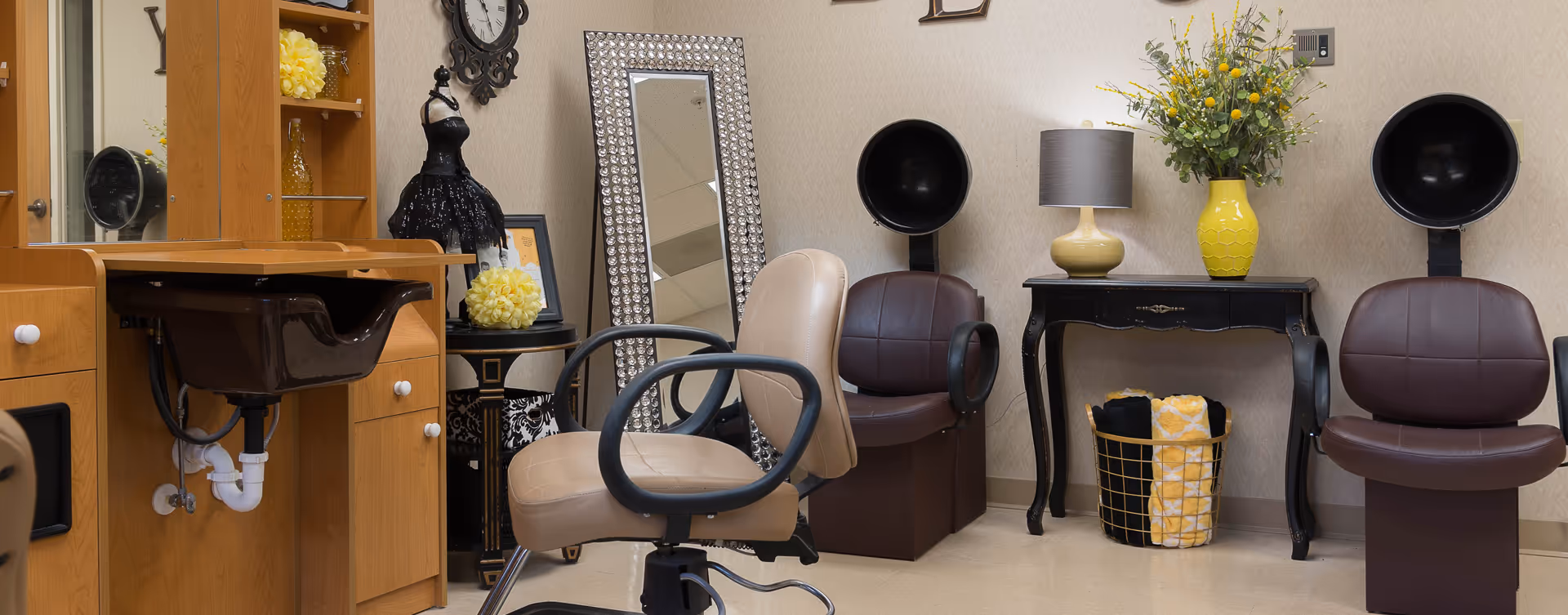 Interior of a salon area with salon chairs, hair drying stations, a wooden vanity with a black sink, a large decorative mirror, a small table with a lamp and a yellow vase with flowers, and a basket with rolled towels.