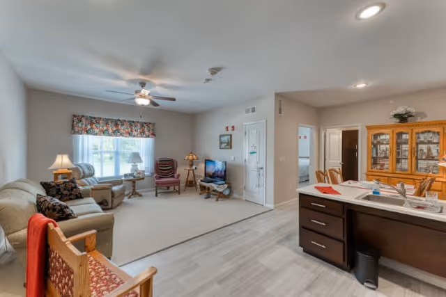 A spacious living area in a senior living facility featuring a beige sofa, armchairs, a wooden chair with a red cushion, a TV on a stand, and a ceiling fan. The room has a large window with floral curtains, a wooden dining hutch, and a kitchen island with a sink and countertop. The flooring is a combination of carpet and light-colored wood.