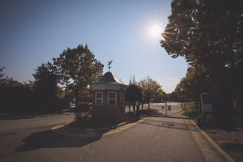 Entrance gate to a facility with a small brick guardhouse topped with a weather vane, surrounded by trees under a clear sky with the sun shining brightly.