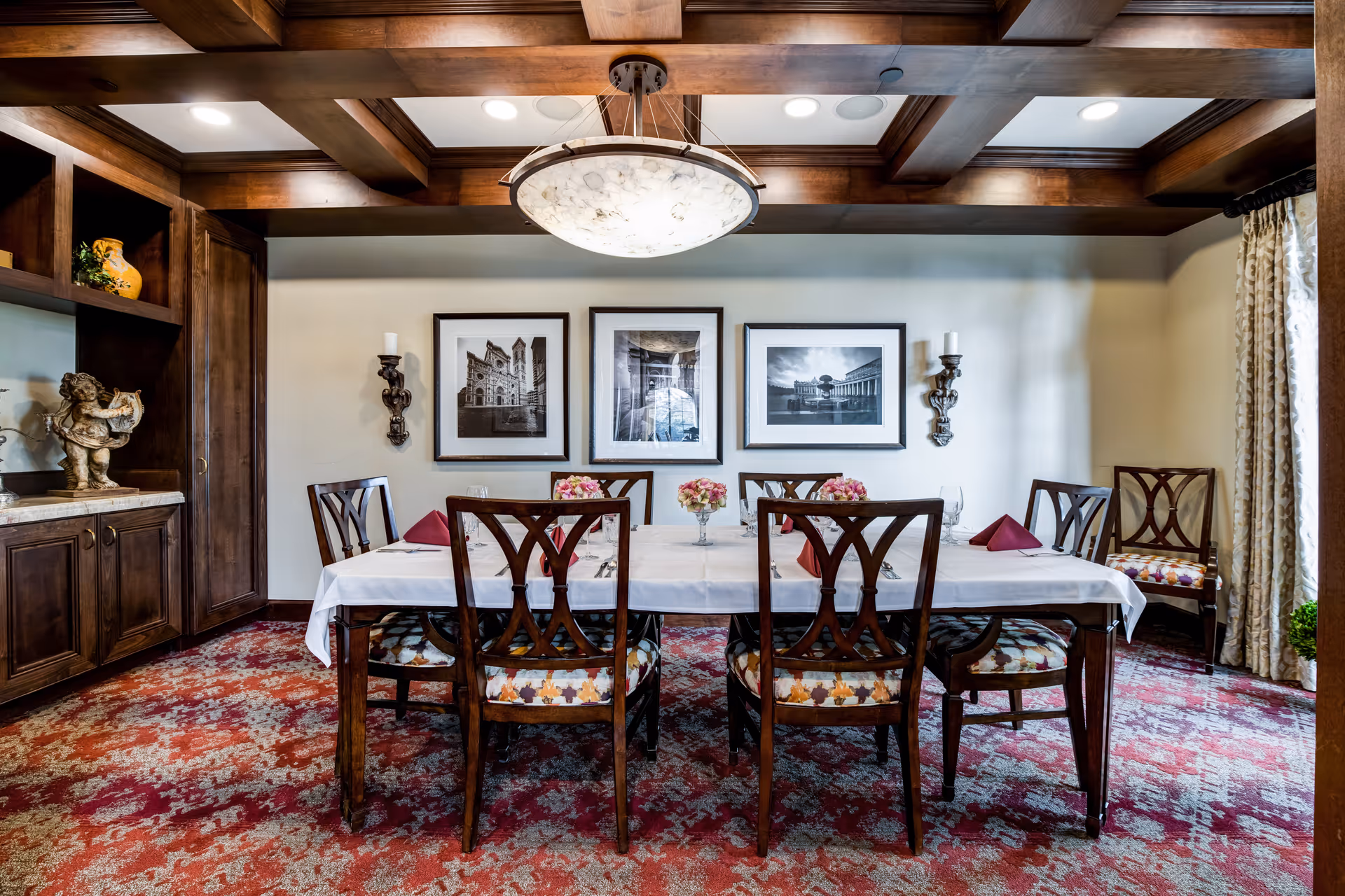 A formal dining room with a rectangular wooden table covered with a white tablecloth and set with glassware, silverware, and burgundy napkins. Six wooden chairs with patterned cushions surround the table. The room features a red and beige patterned carpet, wooden ceiling beams, a large circular chandelier, and three black and white framed photographs on the wall. There are decorative wall sconces with candles and a built-in wooden cabinet with a statue and vase on the left side.