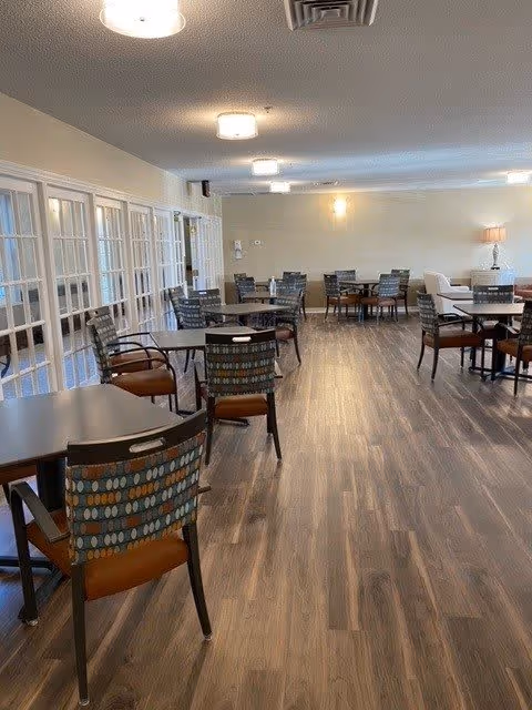 A spacious dining room with multiple tables and chairs arranged neatly on a wood-patterned floor. The room has beige walls, ceiling lights, and large windows with white frames along one side. A lamp is visible on a side table in the background.