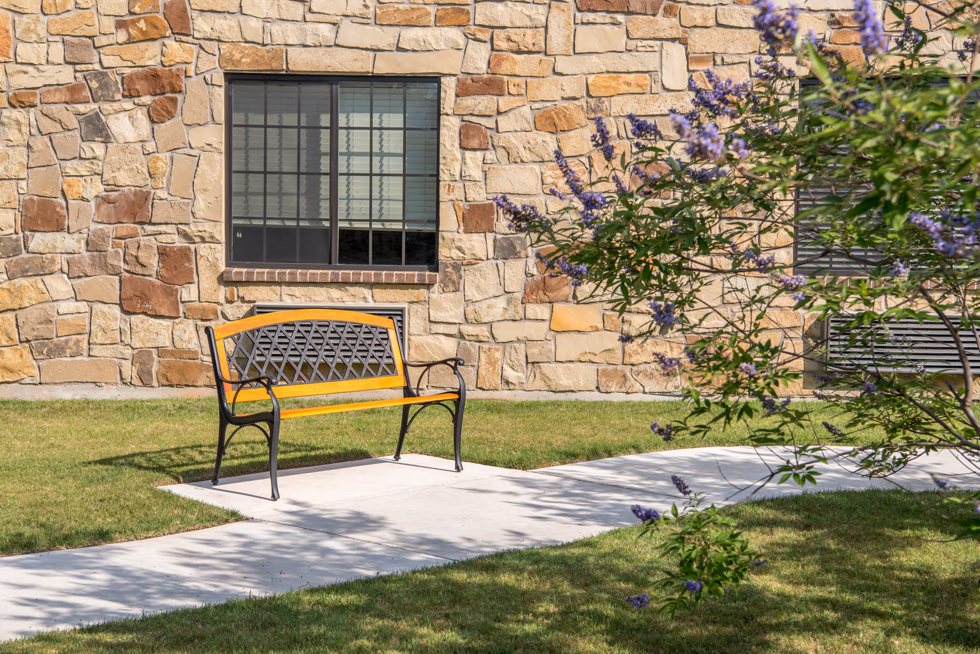 A metal and wood bench sits on a concrete pathway in front of a stone building wall with a window. There is green grass surrounding the pathway and a flowering bush with purple flowers on the right side of the image.