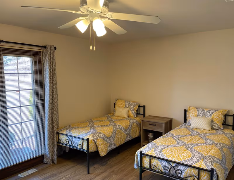 Two twin beds with yellow patterned bedding and a nightstand in a bright bedroom with a ceiling fan and sliding glass door.