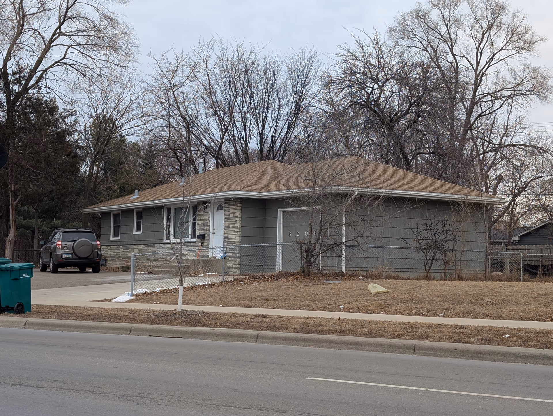 Single-story residential building with a brown roof and gray siding, a white front door, and a garage door with the number 6205. There is a driveway with a parked black SUV, a chain-link fence surrounding the property, leafless trees in the background, and a sidewalk in front of the house.