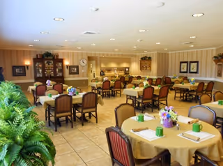 A spacious dining room with multiple tables covered in beige tablecloths, each set with green mugs, napkins, and floral centerpieces. The room has beige tiled floors, striped wallpaper, and recessed ceiling lights. There is a wooden cabinet with glass doors and a clock on the wall, along with framed artwork and plants adding to the decor.