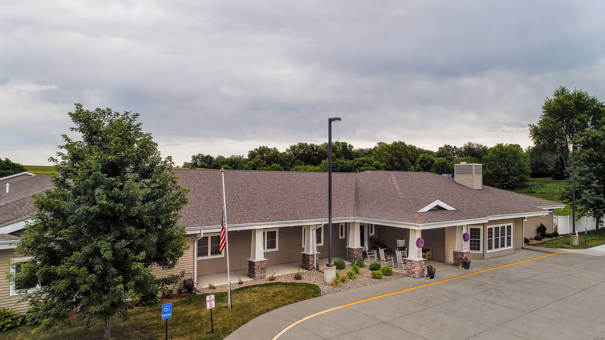 Front entrance of a single-story assisted living building with a covered porch, rocking chairs, an American flag, and a curved driveway.