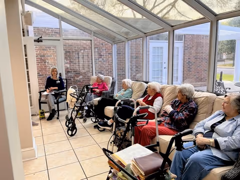 Several elderly residents sitting on a couch and chairs in a sunlit glass-enclosed common room with walkers nearby and a staff member at one end.