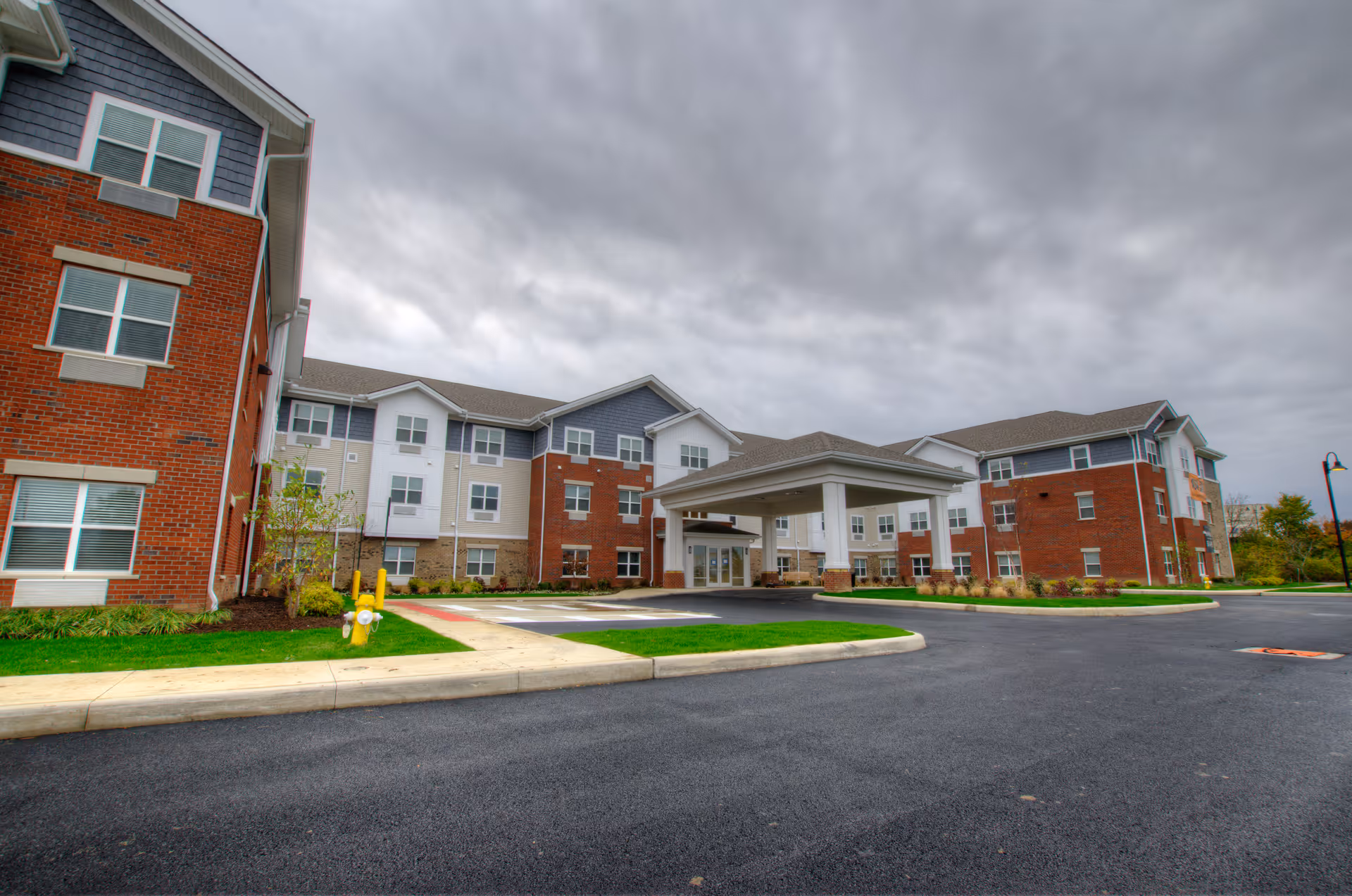 Exterior view of a multi-story senior living facility building with brick and siding facade under a cloudy sky. The entrance features a covered drop-off area with a driveway and landscaped green areas around the building.