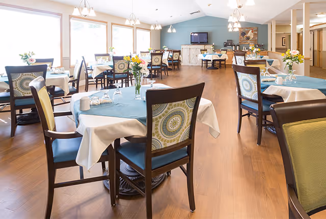 Bright dining room with multiple round tables and upholstered chairs set with tablecloths and flower centerpieces.
