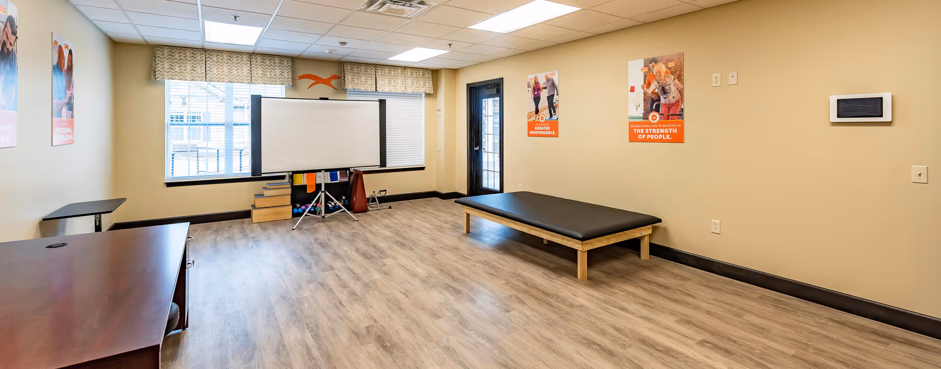 A spacious room with light wood flooring and beige walls, featuring a large window with blinds and a valance, a black padded bench, a projector screen on a tripod stand, a wooden desk, and motivational posters on the walls.