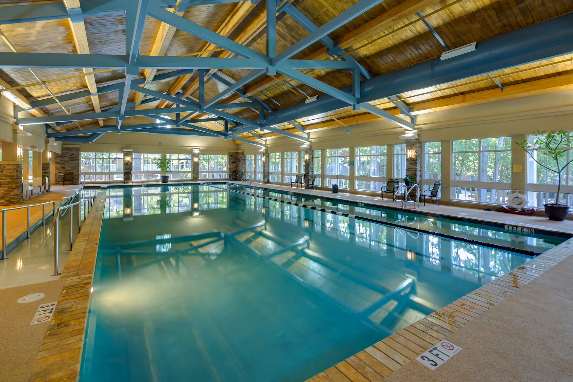 Indoor swimming pool with clear water, surrounded by large windows letting in natural light. The ceiling has exposed wooden beams painted blue, and the pool area includes safety railings and chairs along the sides.