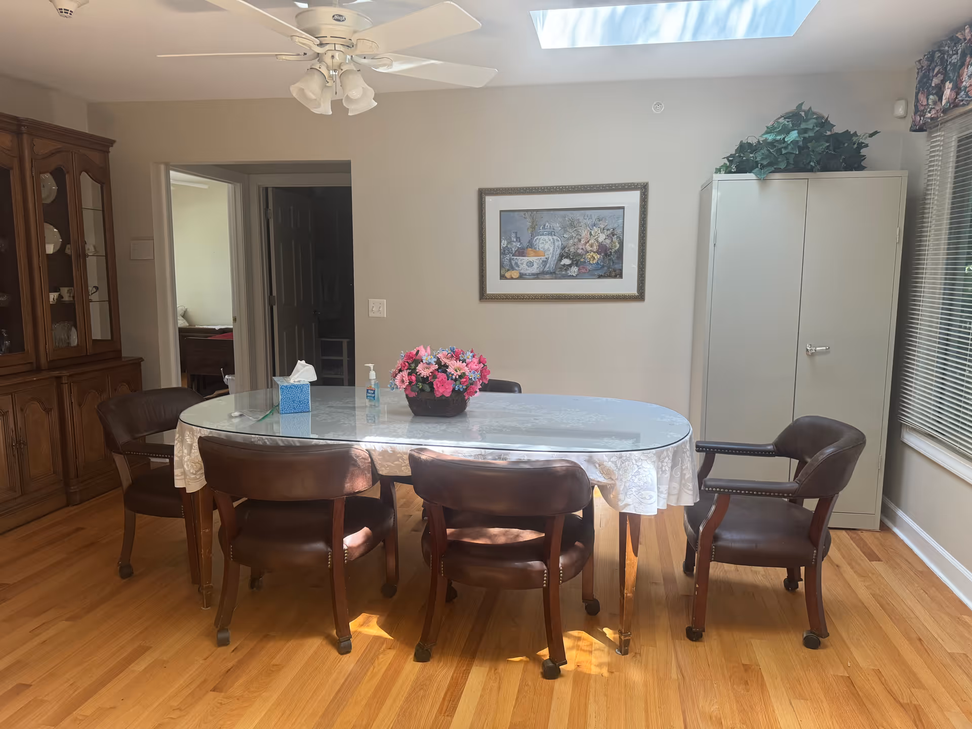 Dining room with oval glass table, six chairs, a wooden cabinet, and skylight.