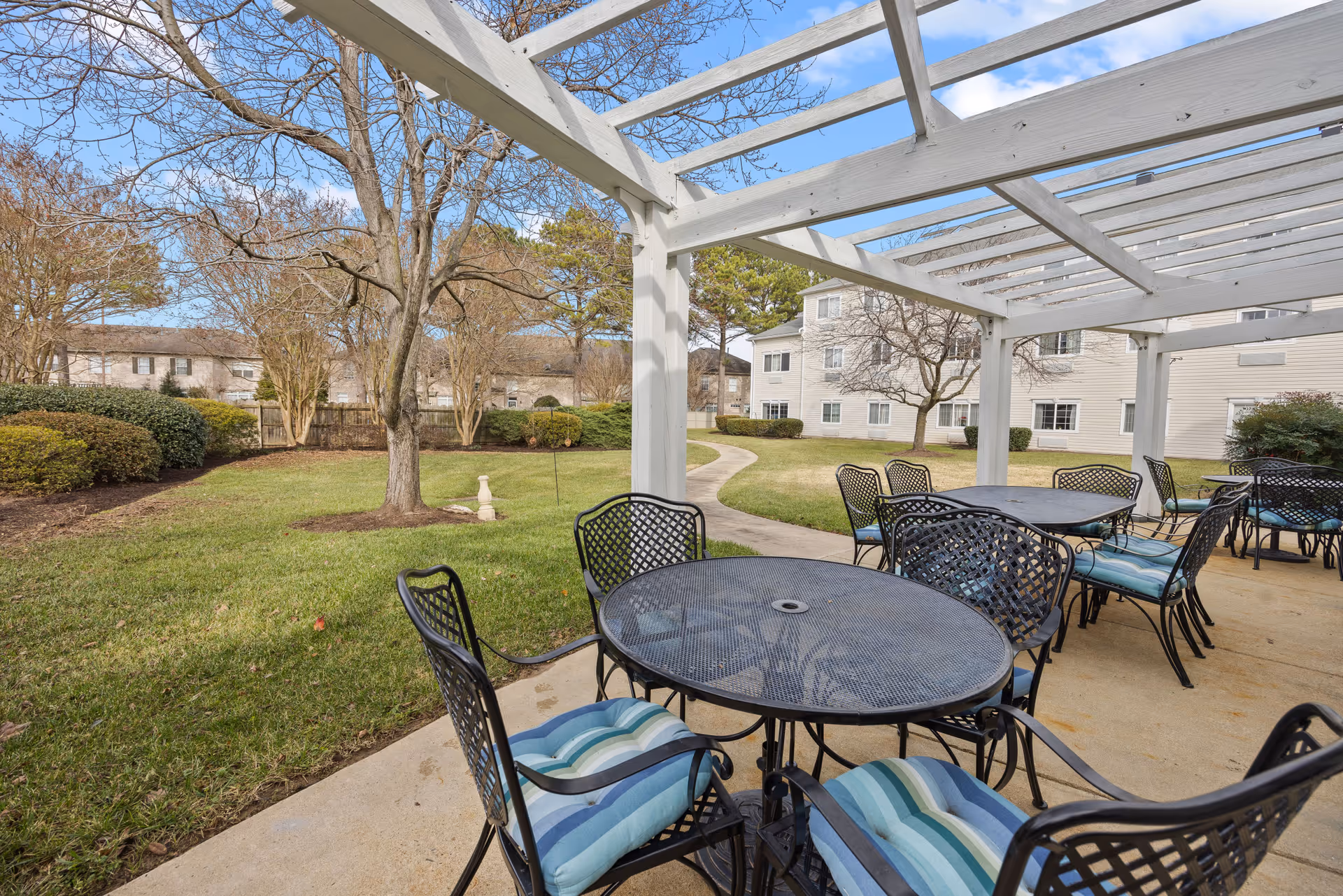 Outdoor covered patio with metal tables and striped-cushioned chairs overlooking a grassy courtyard and residential buildings.