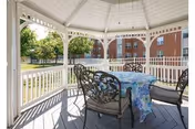 White wooden gazebo furnished with a table covered by a floral tablecloth and metal chairs, overlooking a grassy courtyard and a brick building.