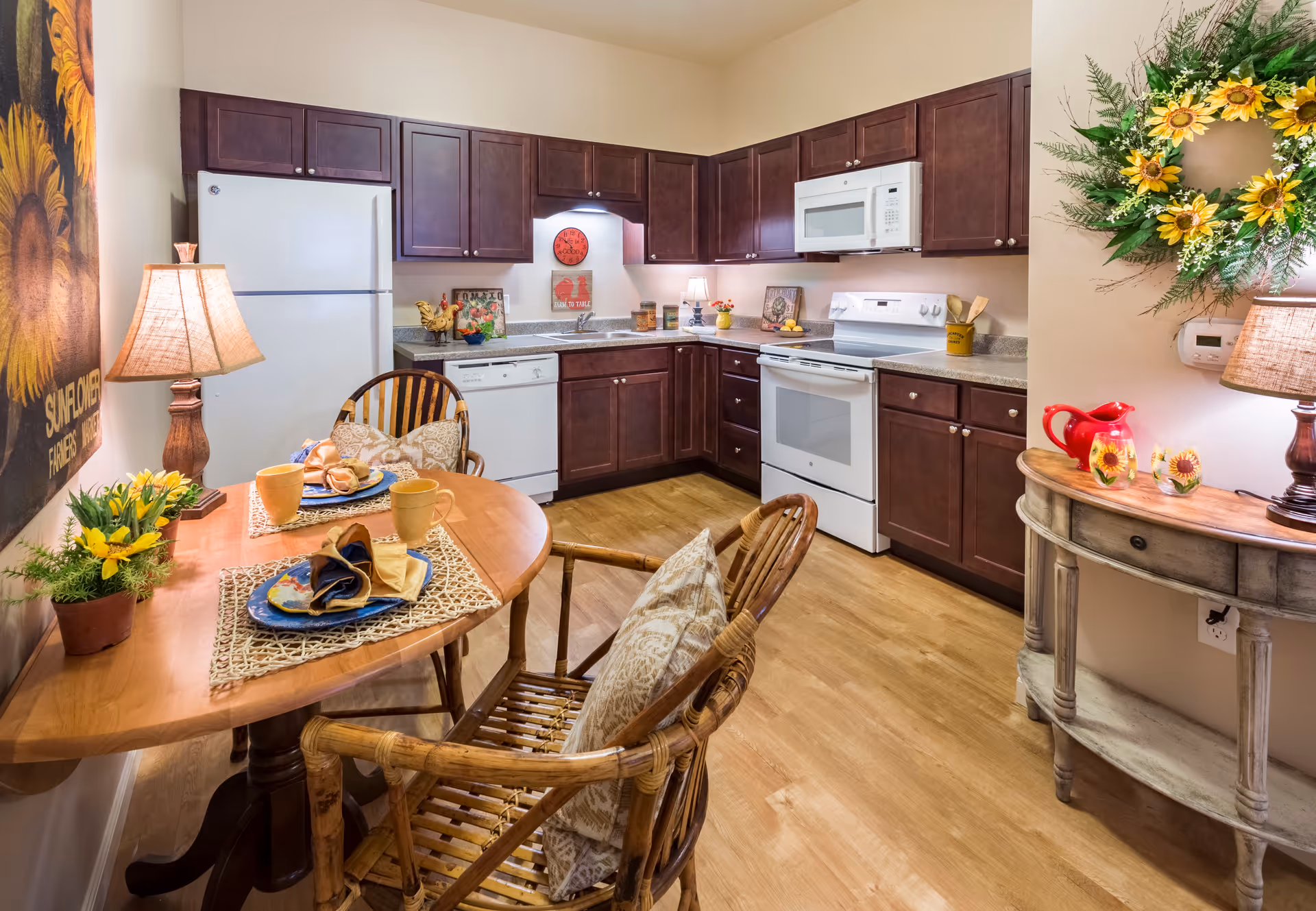A cozy kitchen and dining area featuring dark wood cabinets, white appliances including a refrigerator, dishwasher, stove, and microwave. A small wooden dining table is set with two place settings, yellow mugs, and napkins, accompanied by two wicker chairs with cushions. The room is decorated with a sunflower-themed wall art, a lamp on the table, a small side table with a sunflower wreath above it, and various kitchen decor items.