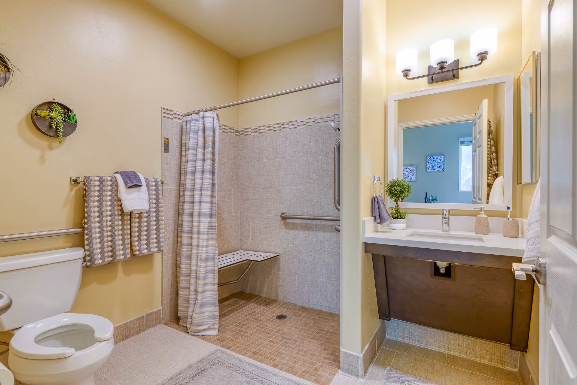 A clean and accessible bathroom with a toilet, a walk-in shower with a fold-down seat and grab bars, a sink with a large mirror above it, and towels hanging on a rack. The walls are painted light yellow and the floor is tiled.