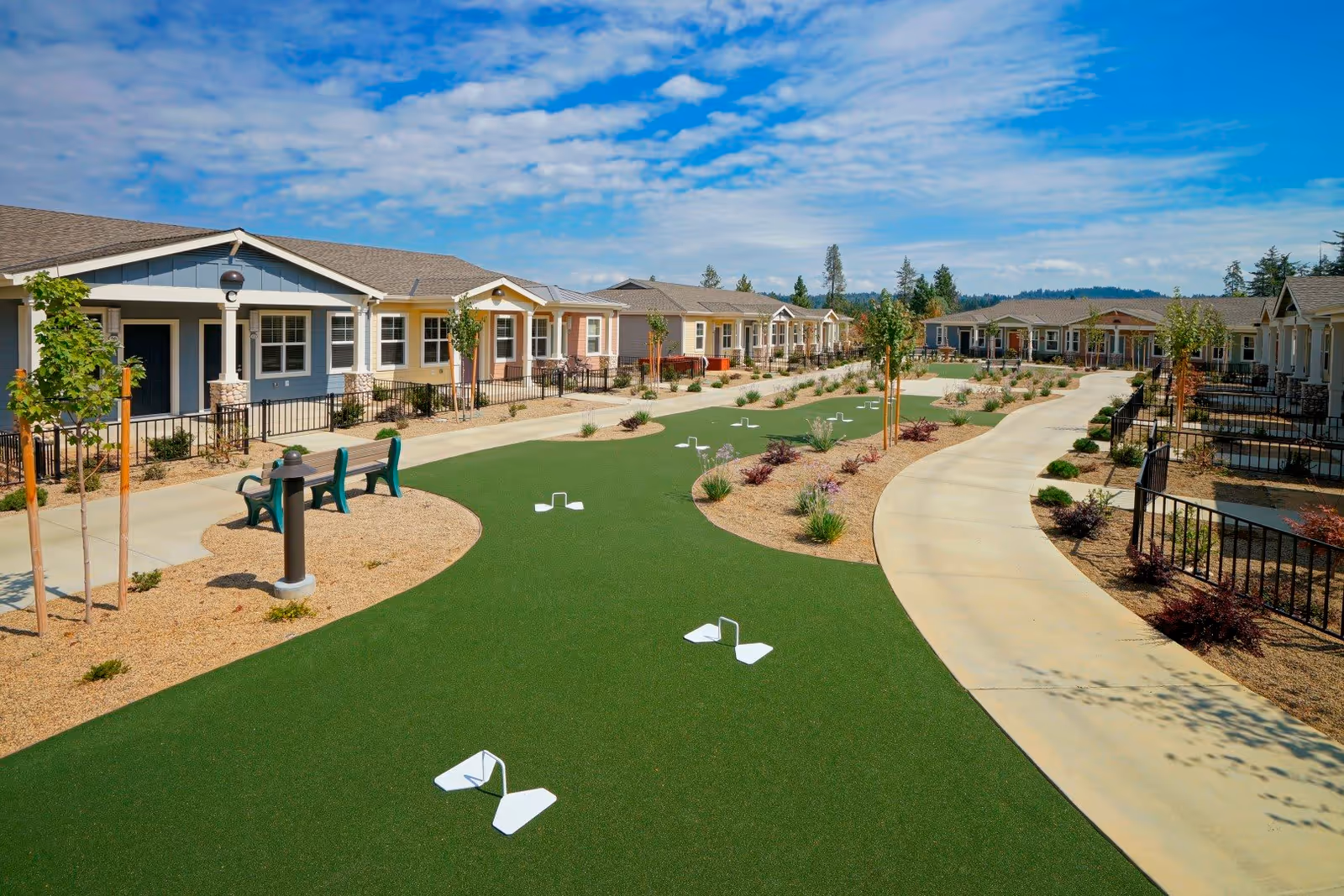 Outdoor view of a senior living community at Eskaton Village Placerville featuring a landscaped courtyard with a putting green, benches, young trees, and a winding concrete pathway. Single-story residential buildings with porches line both sides of the courtyard under a partly cloudy blue sky.