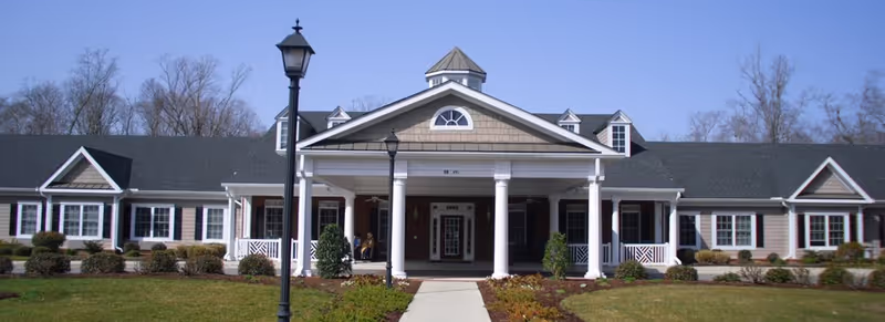 Front exterior view of a single-story senior living facility building with white columns, a covered entrance, multiple windows with black shutters, and a well-maintained lawn and shrubs under a clear blue sky.