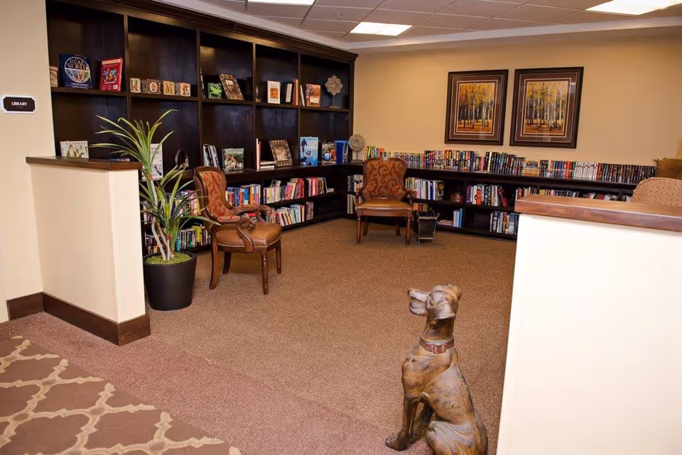 A cozy library room with dark wooden bookshelves filled with books and decorative items. Two upholstered armchairs with patterned fabric are placed near the shelves. A potted plant is positioned near a half wall, and a bronze dog statue sits on the carpeted floor. The walls are painted beige and decorated with two framed pictures of trees.