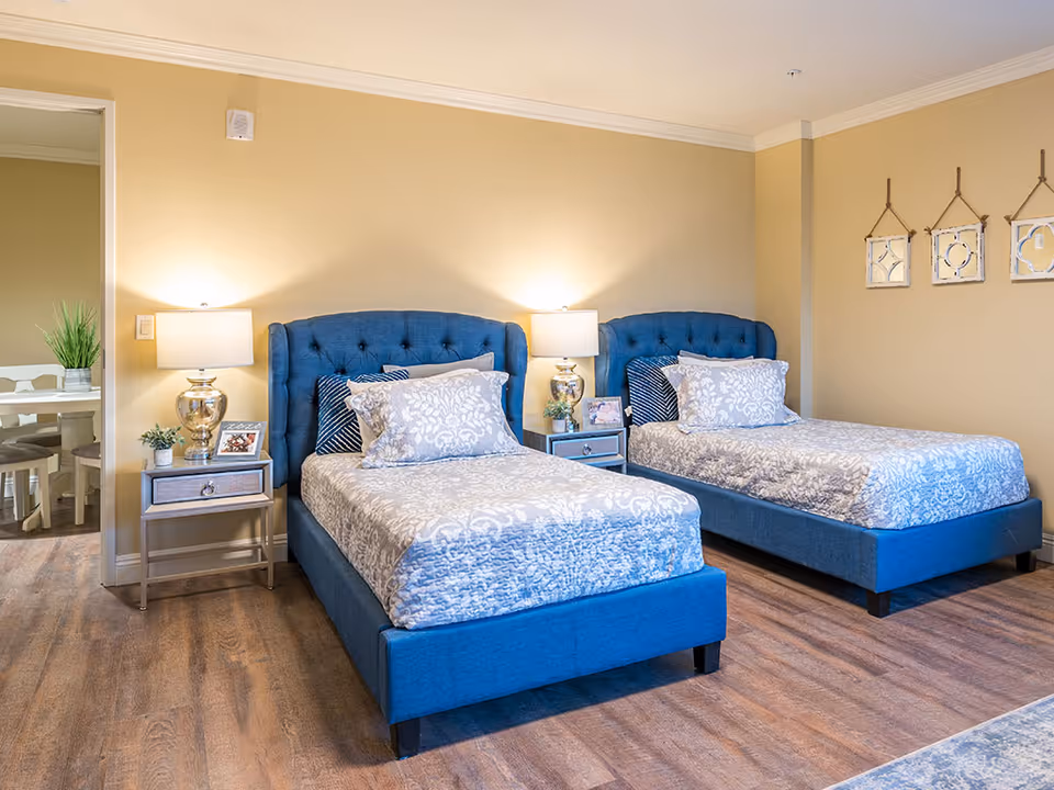 A bedroom with two twin beds featuring blue upholstered headboards and patterned bedding. Each bed has decorative pillows. Between the beds is a nightstand with a lamp, a small plant, and a framed photo. The room has beige walls and wood flooring. On the right wall, three decorative hanging mirrors are displayed. An open doorway on the left leads to a dining area with a white table and chairs.