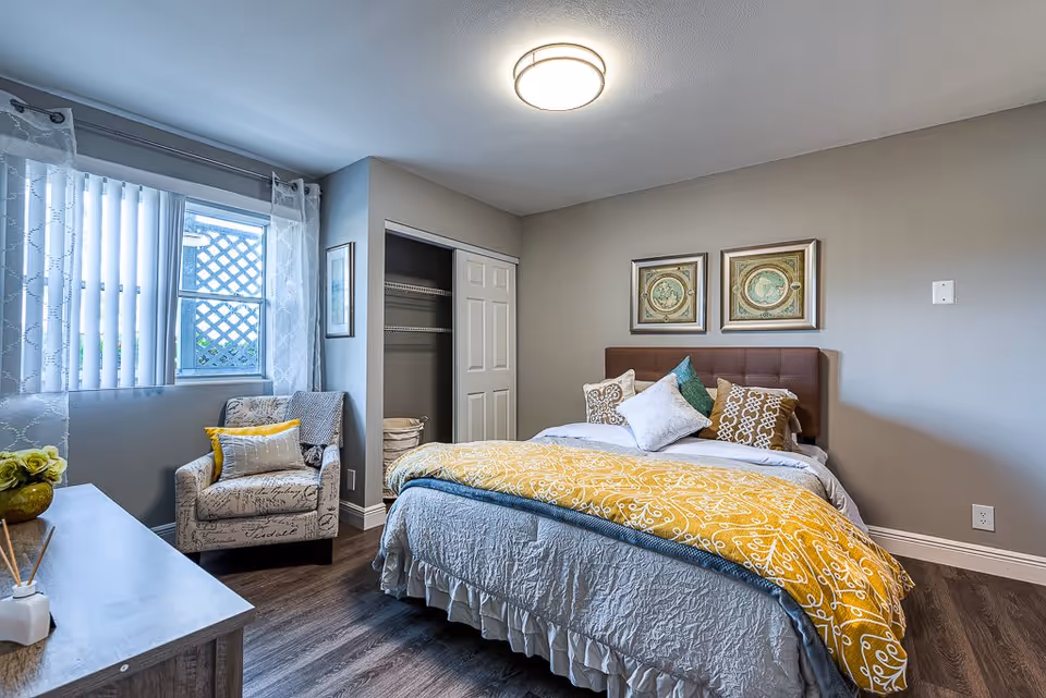 A cozy bedroom with a neatly made bed featuring multiple pillows and a yellow patterned throw blanket. There is a comfortable armchair with a yellow pillow next to a window with sheer curtains and vertical blinds. The room has wooden flooring, a small closet with sliding doors, two framed artworks above the bed, and neutral-colored walls.