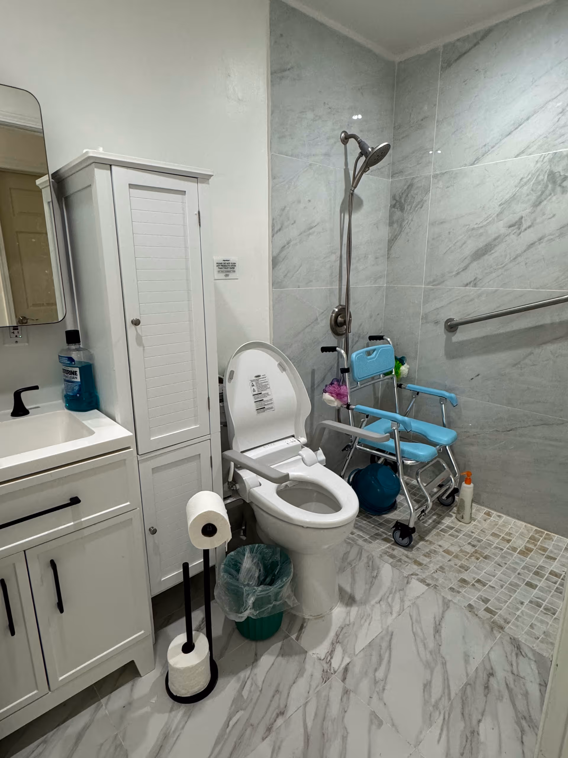 A bathroom with a white toilet equipped with armrests, a white cabinet, a sink with a black faucet, a toilet paper holder with two rolls, a green trash bin, a blue shower chair with wheels, a handheld showerhead, and a grab bar on the tiled wall. The floor and walls have a marble-like design.