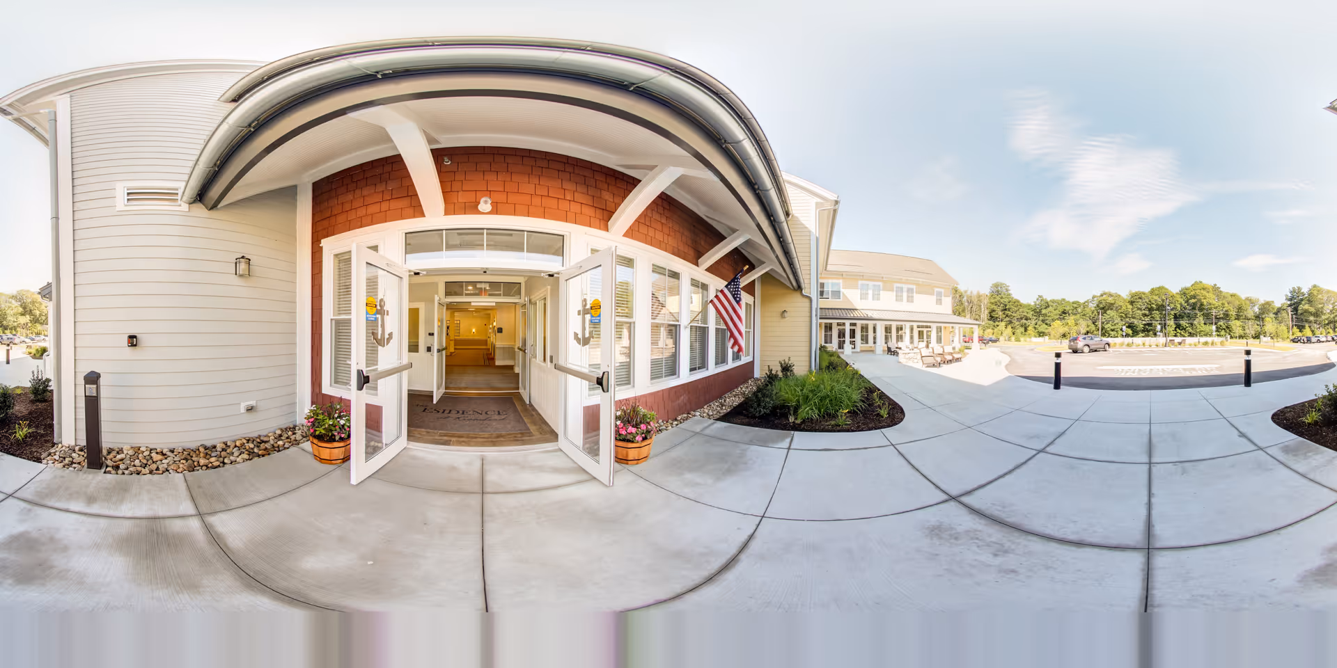 Front entrance of a senior living residence with double doors open under a covered entry, planters, an American flag, and a circular driveway.