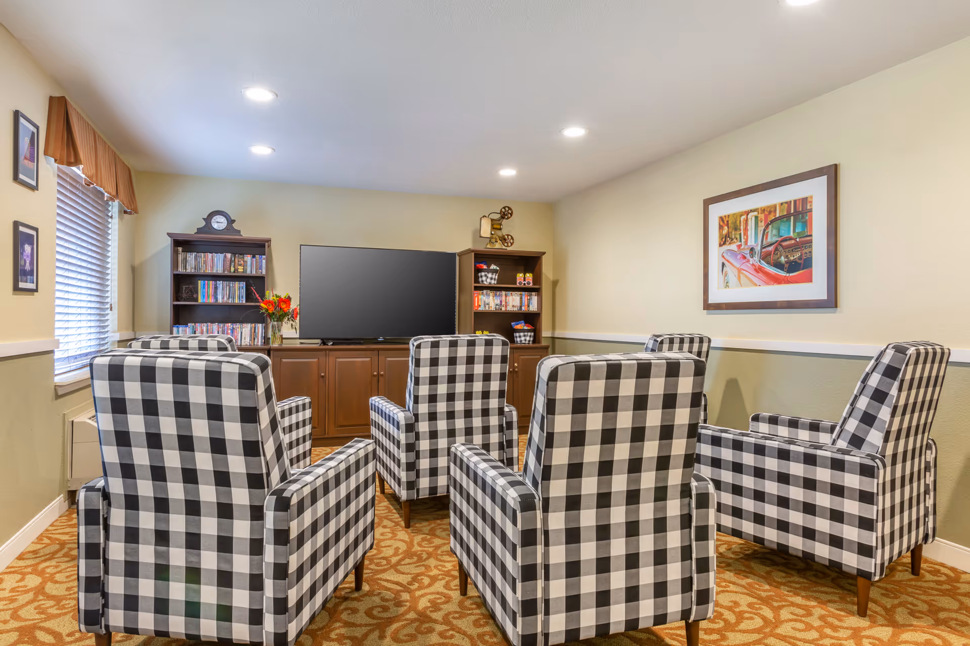 A cozy living room with five black and white checkered armchairs arranged facing a large flat-screen TV on a wooden cabinet. The room has beige walls with a green lower half, a patterned carpet, a window with blinds and a valance, bookshelves filled with DVDs and decor items, and a framed picture of a red vintage car on the wall.